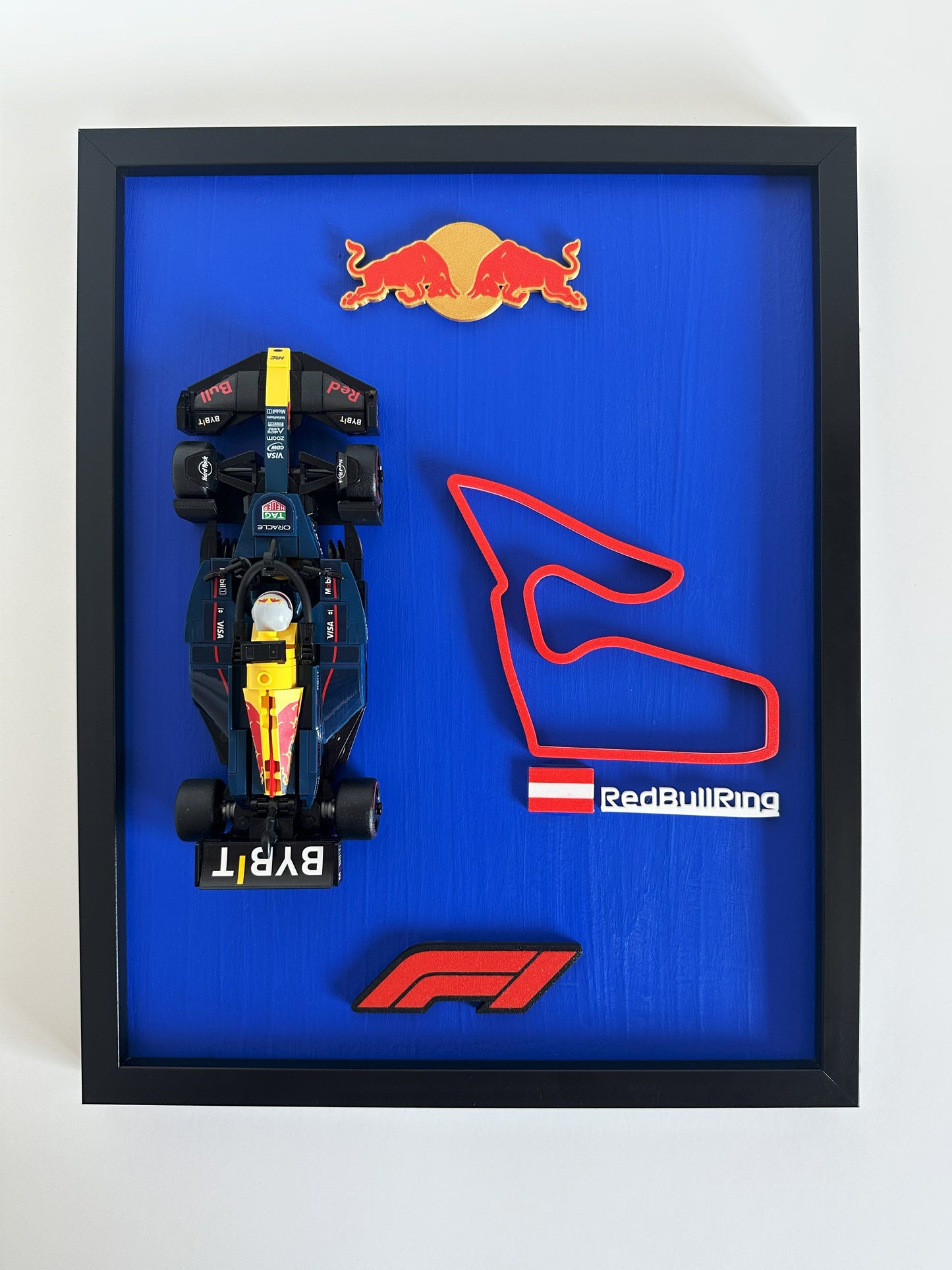Framed display of a Formula 1 car model and Red Bull Ring track layout with Red Bull logo on a blue background.